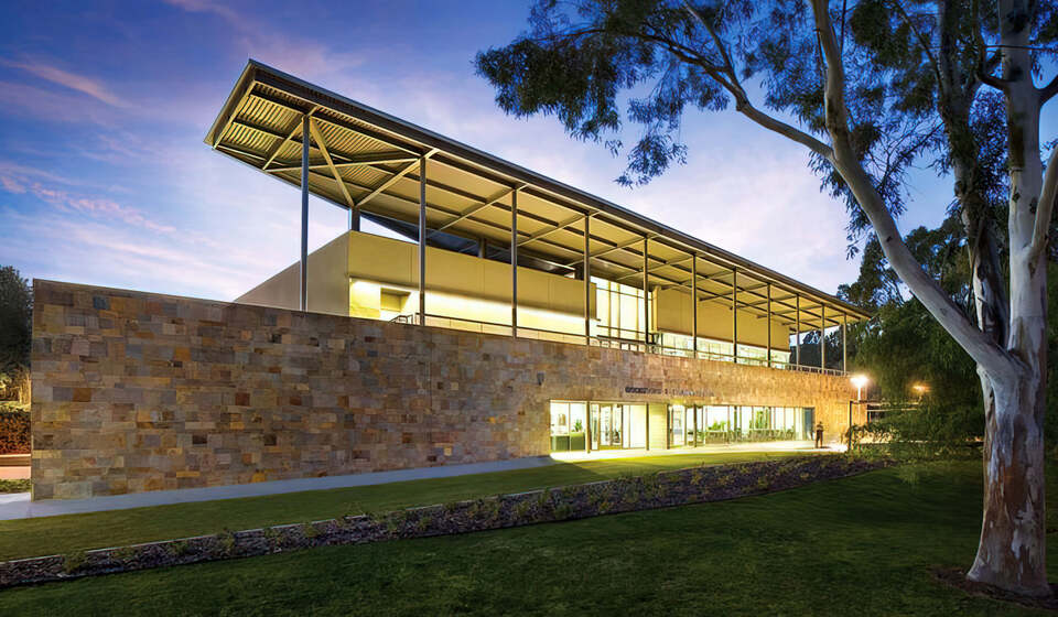 The Santiago Canyon College Learning Resource Center in Orange, California, a modern two-story building with large glass windows and stone walls, illuminated at twilight, surrounded by trees. By LPA Design Studios.